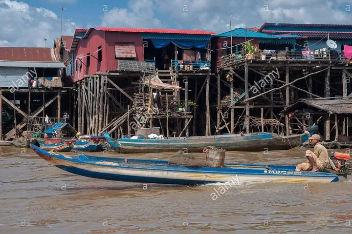 A man piloting a longtail boat past stilted houses on the river.