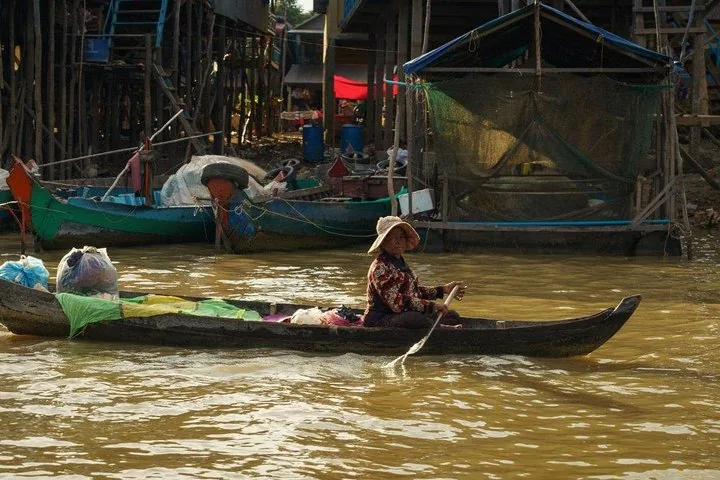 Eine Frau mit Hut rudert ein kleines Holzboot in der Nähe von Pfahlbauten.